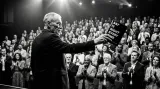 Eamon Blackthorn raising Say It Right Every Time toward a standing, applauding auditorium under stage lighting